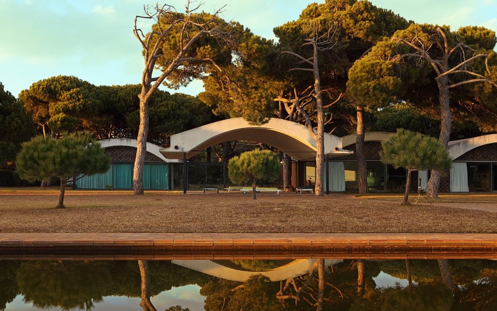 Exterior of Casa Gomis with curving arches, reflective pond, and serene pine tree surroundings at sunset.