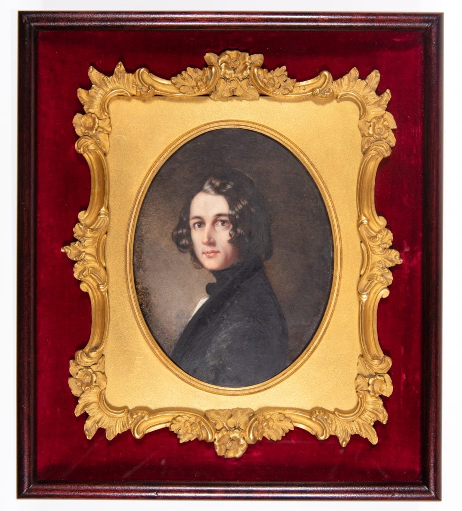 a small oval painted portrait of a young man in profile turning to look at us and wearing black, there is an elaborate square gold frame