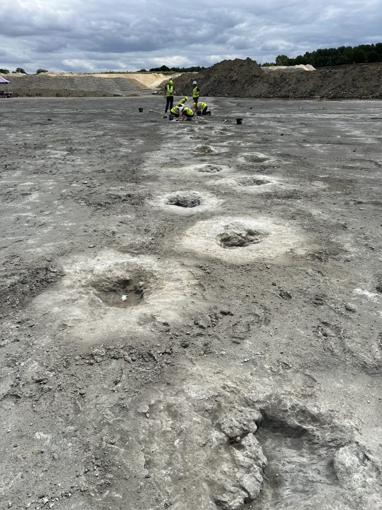 A series of preserved dinosaur footprints in a U.K. quarry, with workers conducting an excavation in the background.