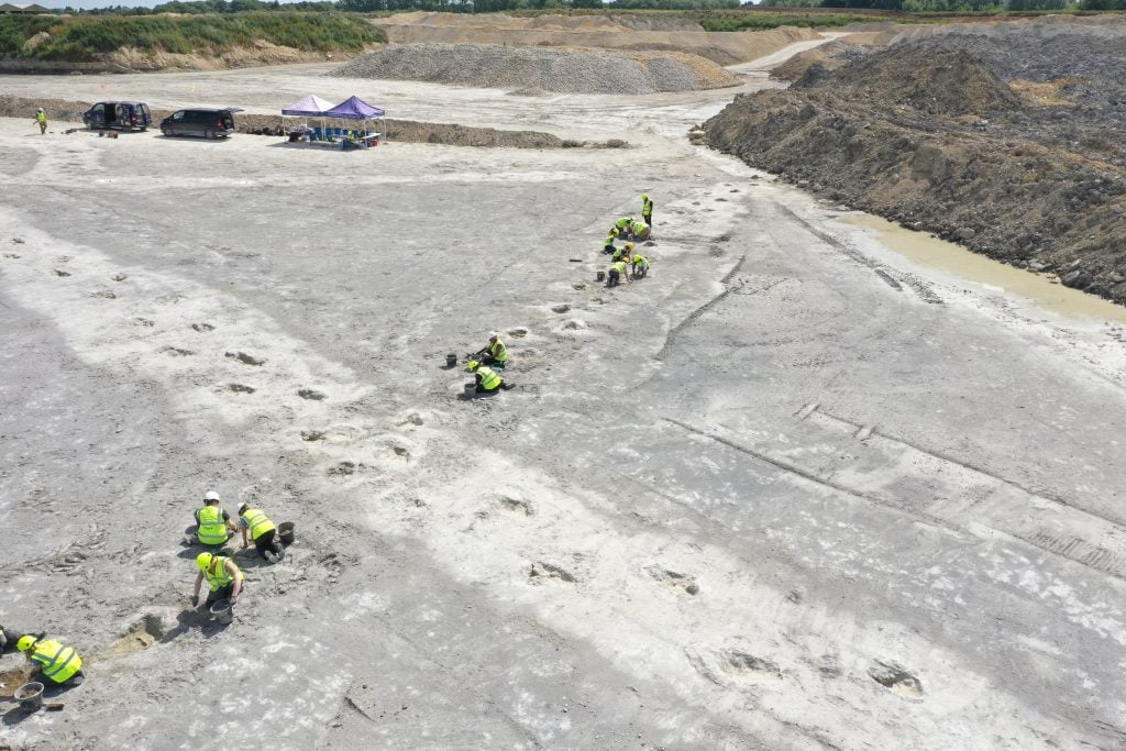 Wide view of a U.K. quarry excavation site, showing a row of dinosaur footprints and workers.