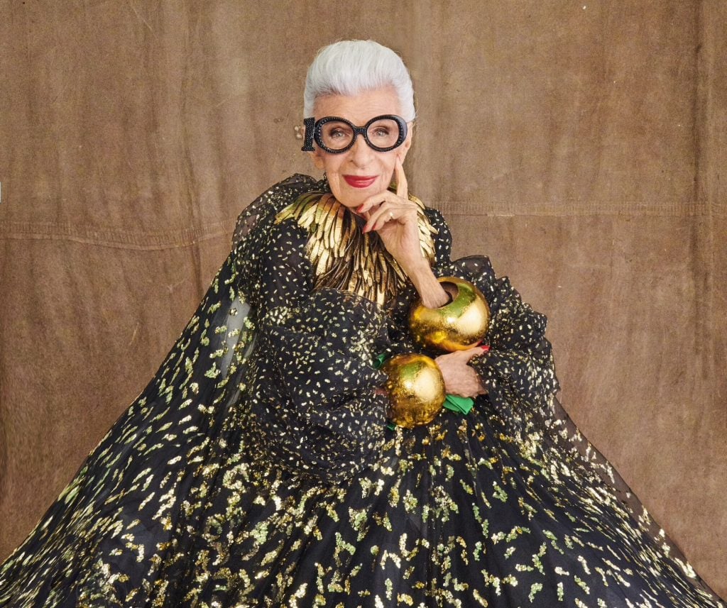 An elegant portrait of Iris Apfel in a black-and-gold gown, accessorized with bold glasses and jewelry.