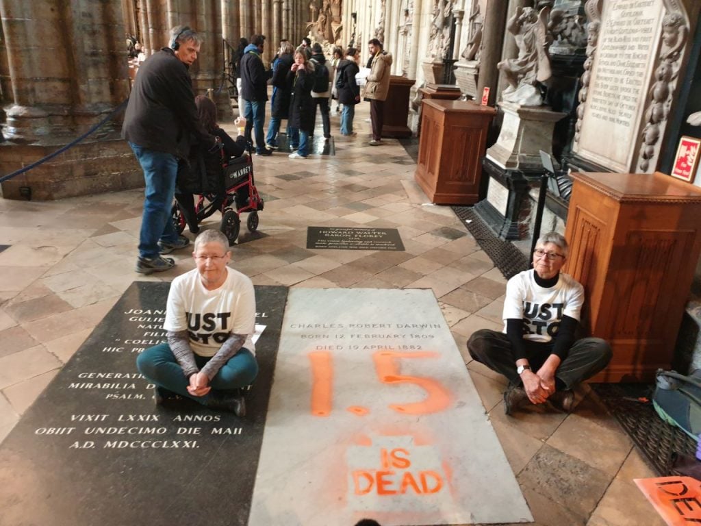 two people sit on a marble floor in front of a grave stone and the grave stone bears the orange graffitied words '1.5 is dead'