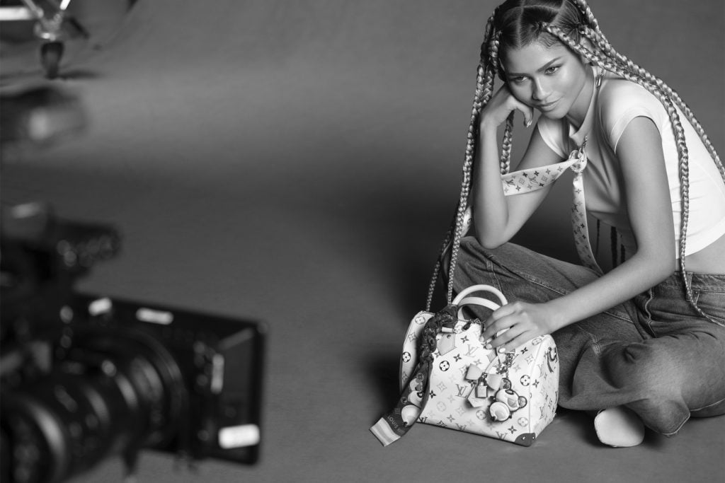 Zendaya sitting cross legged at a photo shoot, with a Louis Vuitton purse in front of her