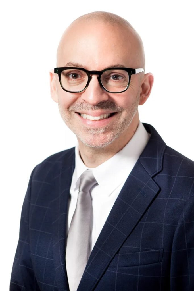 headshot of a man in black-rimmed glasses and a suit 