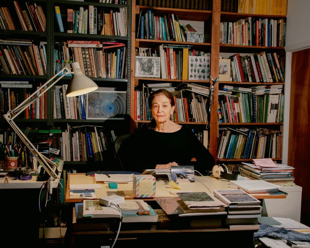 Artist Mary Miss seated behind a desk in a room lined with filled bookshelves.