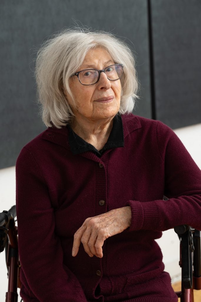 A color portrait of an older woman with glasses and a maroon shirt with her arm folded in front of her body.