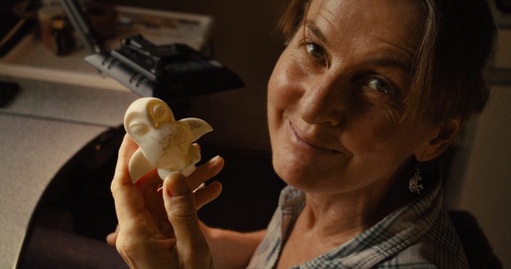 An artist looks at the camera with a smile, holding a figurine of an owl