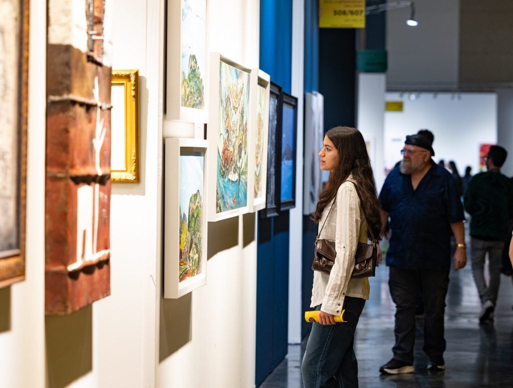 A visitor at Art Palm Beach looking at a wall covered in framed artworks.