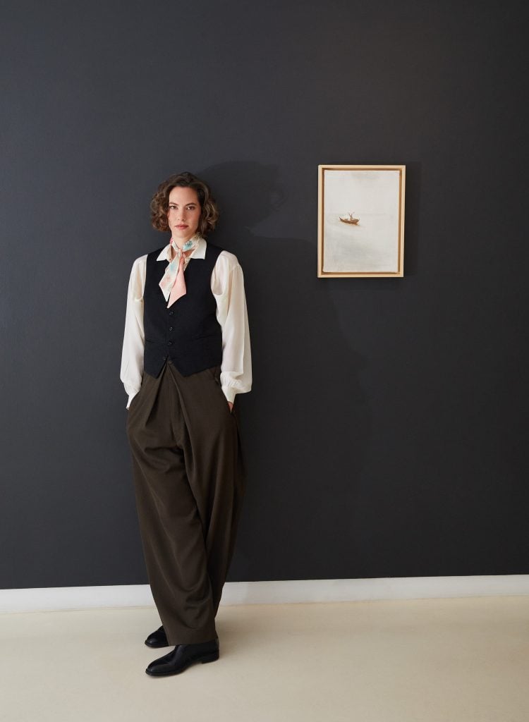 Emma Ferrer posing in a white shirt and black vest next to her painting of a lamb floating in a sea