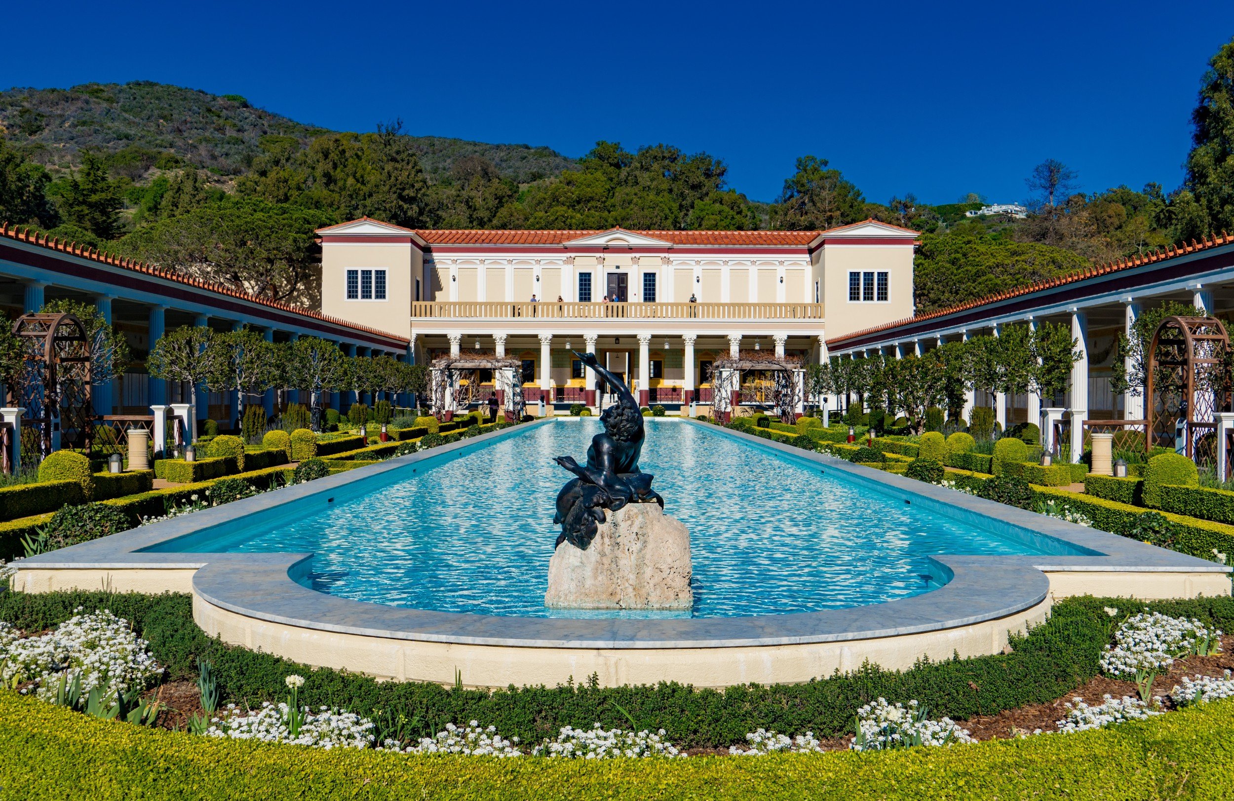 Getty Villa courtyard with reflective pool, statue centerpiece, manicured gardens, and Mediterranean-style architecture under clear skies.