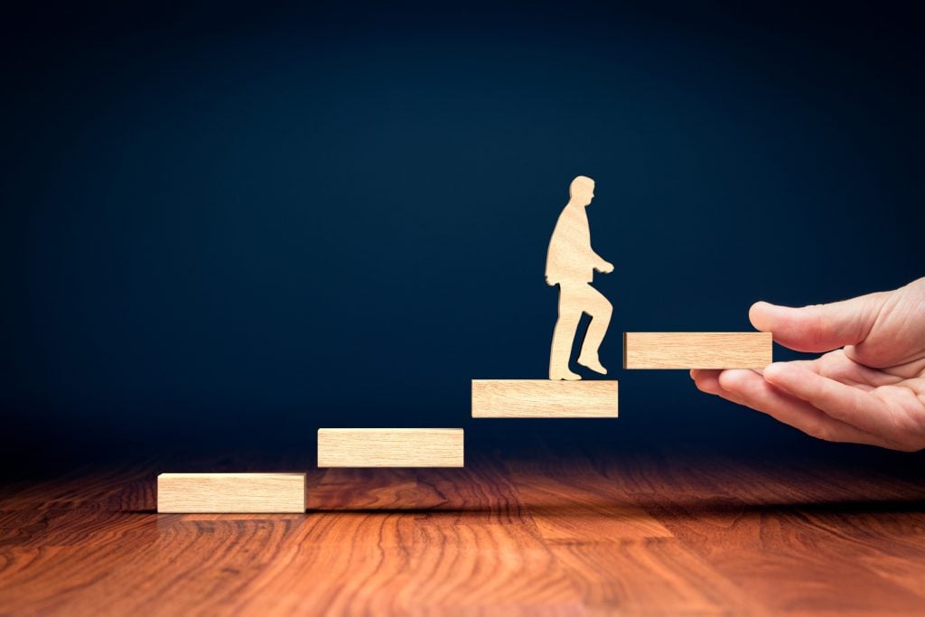 A wooden cutout of a person climbs steps as a hand supports progress, symbolizing mentorship.