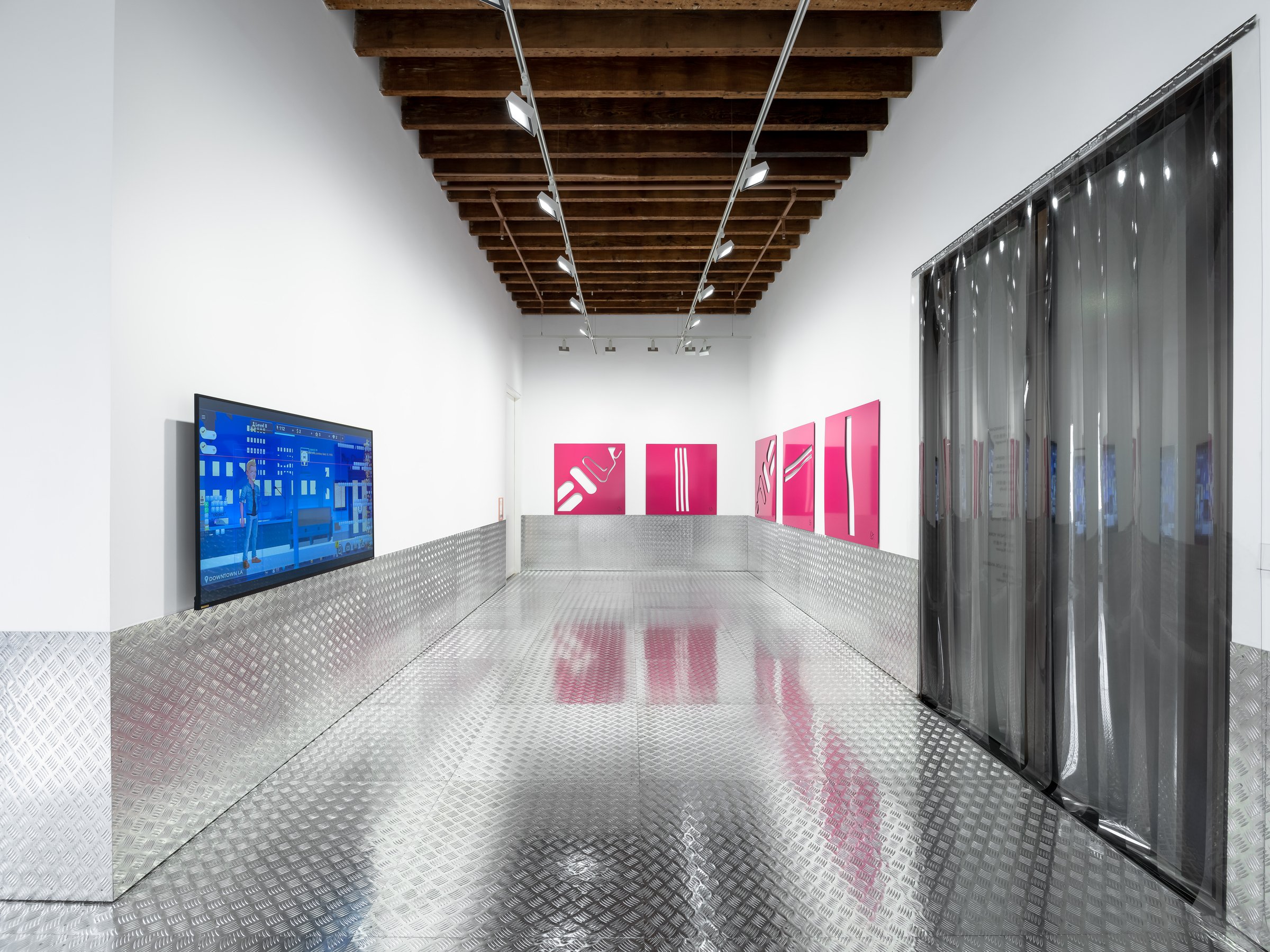 A white-walled gallery holds a screen with a video game and a number of rectangular pink pictures