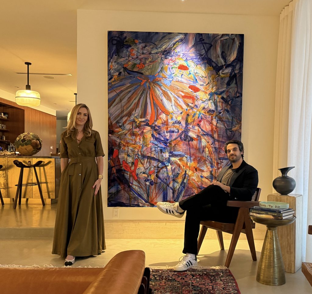 A stylishly dressed woman and man pose in a modern living space with a large, colorful abstract painting on the wall.