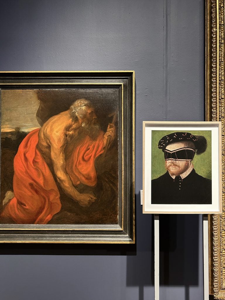 A museum display featuring a classical painting of an elderly man in red robes alongside a modern artwork depicting a historical portrait with a surreal blindfold.