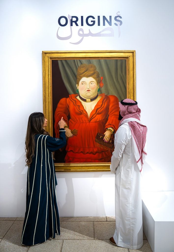Two people in traditional attire admire a large, framed painting of a woman in a red dress at an art exhibition titled "ORIGINS."