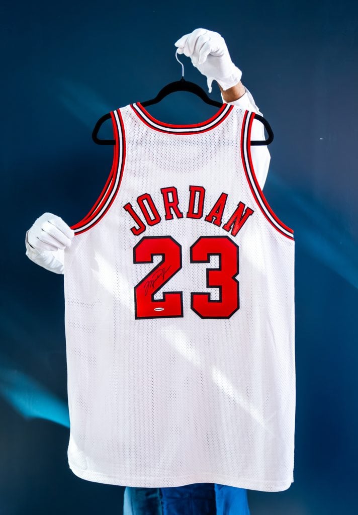 Two white-gloved hands hold a white basketball jersey from behind, that reads jordan 23 in red letters