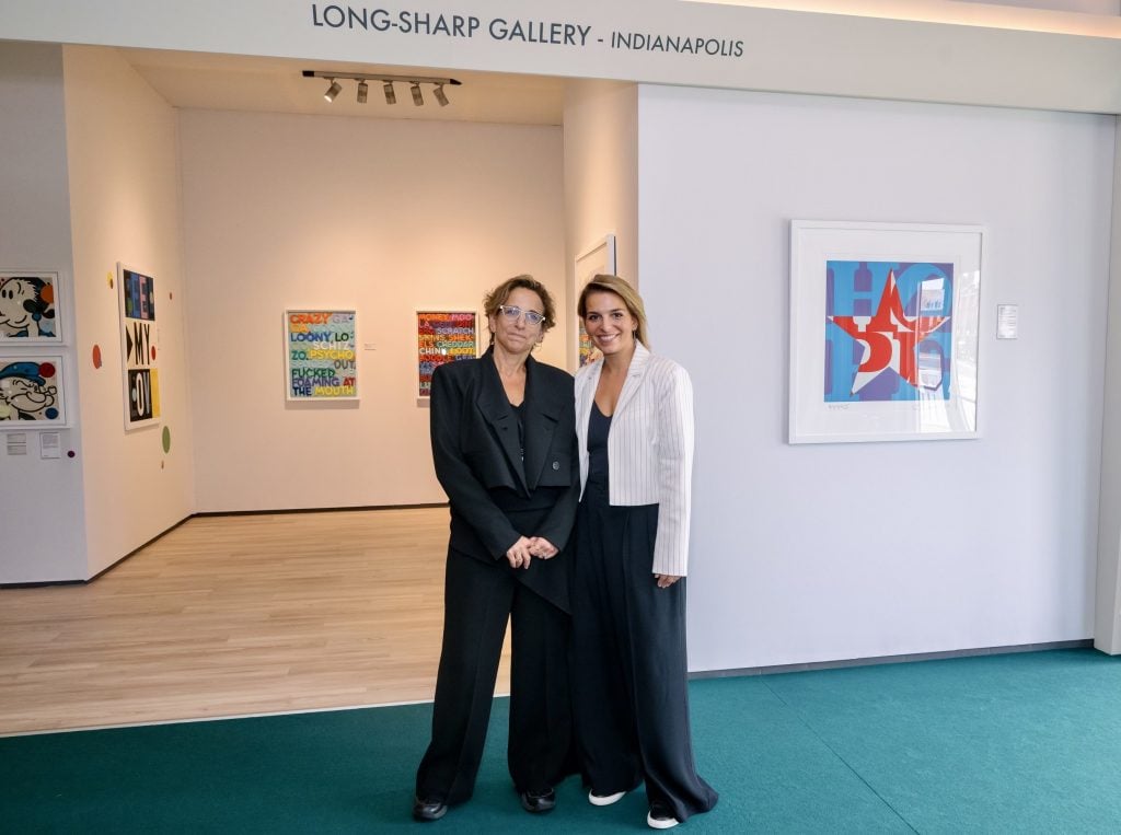 Two women stand side by side in front of the entrance to Long-Sharp Gallery in Indianapolis. They are dressed in black outfits with light jackets and are smiling at the camera. Inside the gallery, colorful contemporary artworks, including by Andy Warhol, hang on the walls, including text-based and pop art-inspired pieces. A framed artwork with a red star and blue background is visible on the right wall.