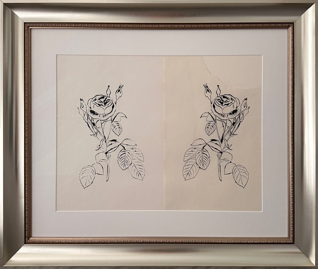 Framed artwork featuring two mirrored black ink drawings of a single rose stem with leaves and buds, executed in Andy Warhol's signature "blotted line" technique. The delicate lines and slight asymmetries reflect the print-transfer process used to duplicate the image. Displayed side by side on cream paper within a wide white mat and silver-toned frame.