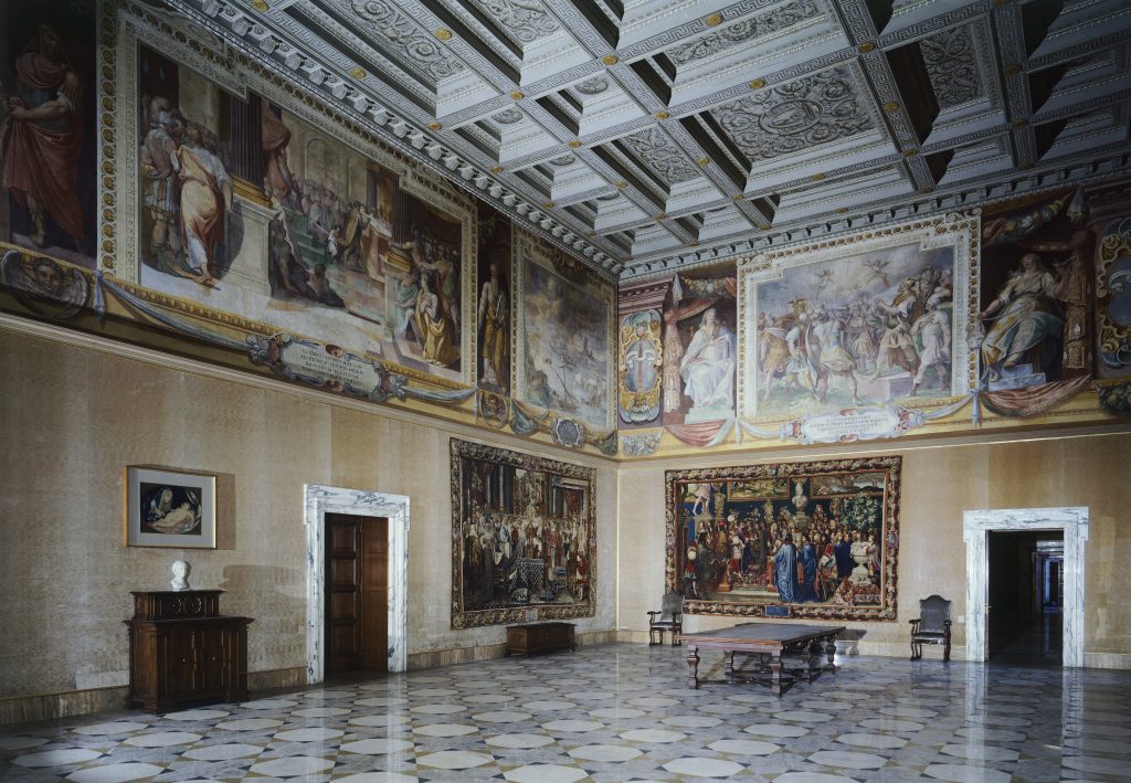 a large stately room that contains frescoes near the ceiling and other works of art hanging on the wall below