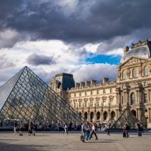 A photo of the Louvre Museum in Paris featuring the large glass pyramid entrance in the courtyard, with people walking and gathering under a dramatic, cloudy sky.
