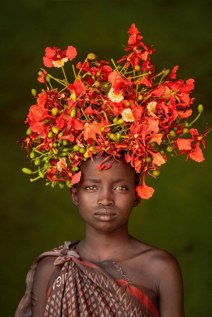 A striking portrait by Cristina Mittermeier titled Wild Flowers in My Hair (2023), depicting a young person from Ethiopia’s Omo Valley wearing an elaborate headdress made of vibrant red and orange flowers with green buds. The subject gazes directly at the camera with a calm, dignified expression, set against a soft green background that enhances the vivid colors of the floral arrangement.