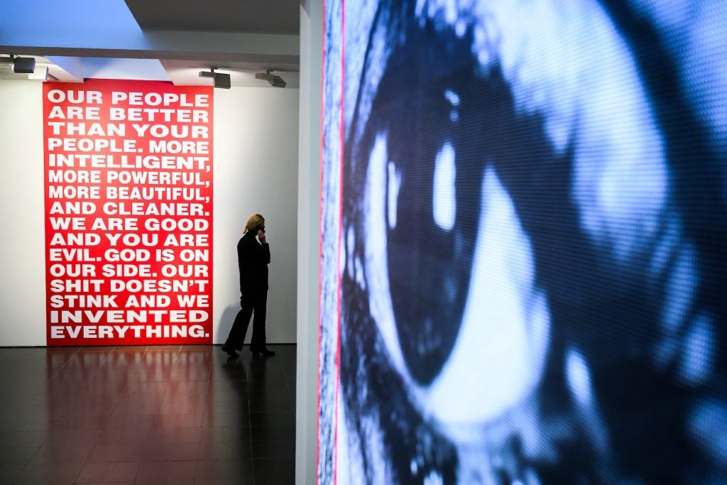 A gallery visitor walks past two large artworks. On the left, a bold red sign with white text reads, “OUR PEOPLE ARE BETTER THAN YOUR PEOPLE. MORE INTELLIGENT, MORE POWERFUL, MORE BEAUTIFUL, AND CLEANER. WE ARE GOOD AND YOU ARE EVIL. GOD IS ON OUR SIDE. OUR SHIT DOESN’T STINK AND WE INVENTED EVERYTHING.” On the right, a large screen displays a fragmented, pixelated close-up of a human eye.