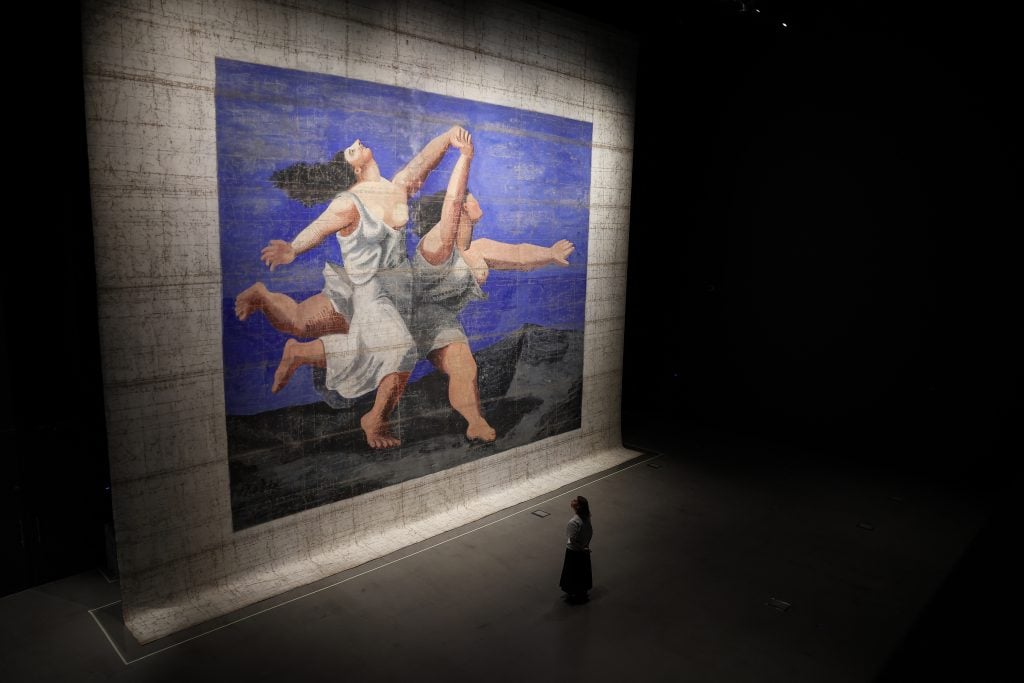 Large stage backdrop painted by Pablo Picasso for Le Train Bleu, showing two women in white dresses dancing against a vivid blue background, with a person standing in front for scale.