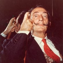 Salvador Dalí in suit with red tie stares wide-eyed, holding cane beside curled mustache dramatically.