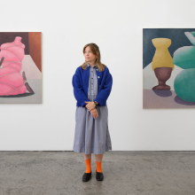 A woman in a blue sweater stands in front of a colorful abstract painting with large orange shapes.