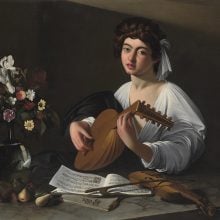 a painted scene in which a young boy plays a lute, he wears a loose and low-cut white shirt and he is at a desk with a vase of flowers, some fruit, and an open book of music