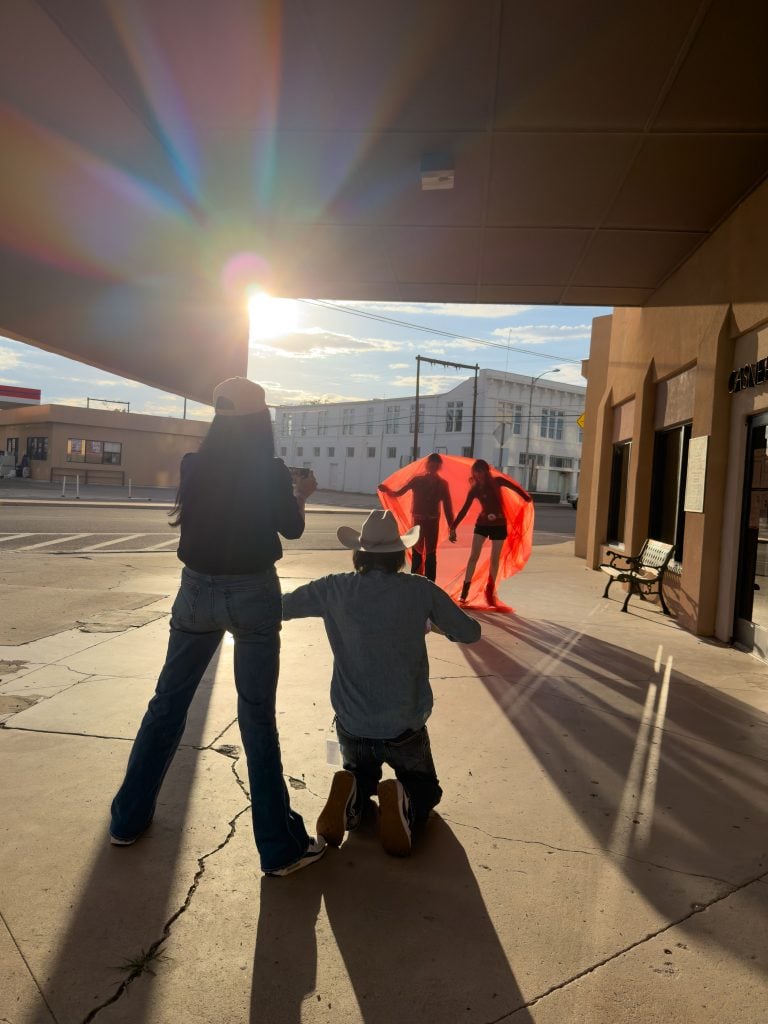 Behind-the-scenes photo of two figures under red fabric, filmed by crew in afternoon sunlight.