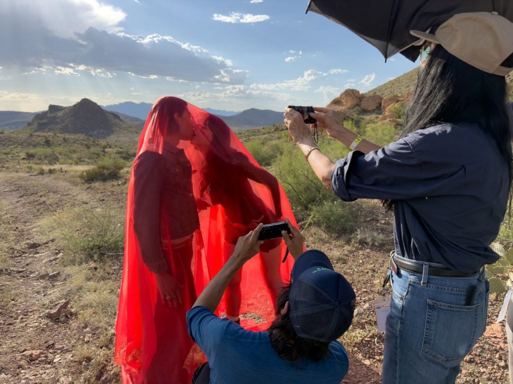 Two figures kiss under red fabric in desert, filmed by crew using cameras and umbrella.
