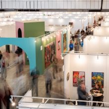 Wide view of bustling art fair with white booths, colored partitions, lights, and blurred visitors moving through aisles; artworks line walls; conversations occur at tables.