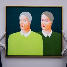 Two attendants in white shirts and blue aprons hold up a painting of two men, one in bright green and one in dark green shirts