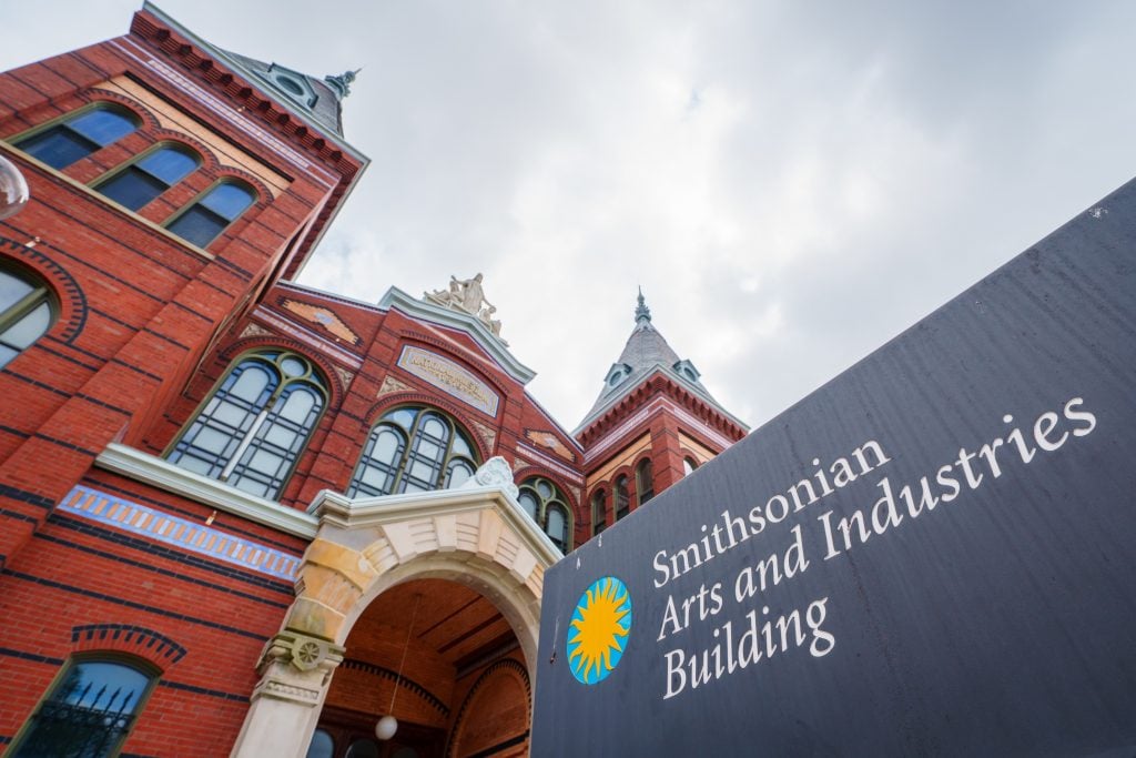 Exterior view of the Smithsonian Arts and Industries Building in Washington, D.C., featuring its historic red-brick architecture with towers and arched windows alongside the Smithsonian signage.