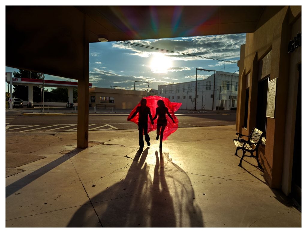Two silhouetted figures run under red fabric, casting long shadows on sunlit city sidewalk.