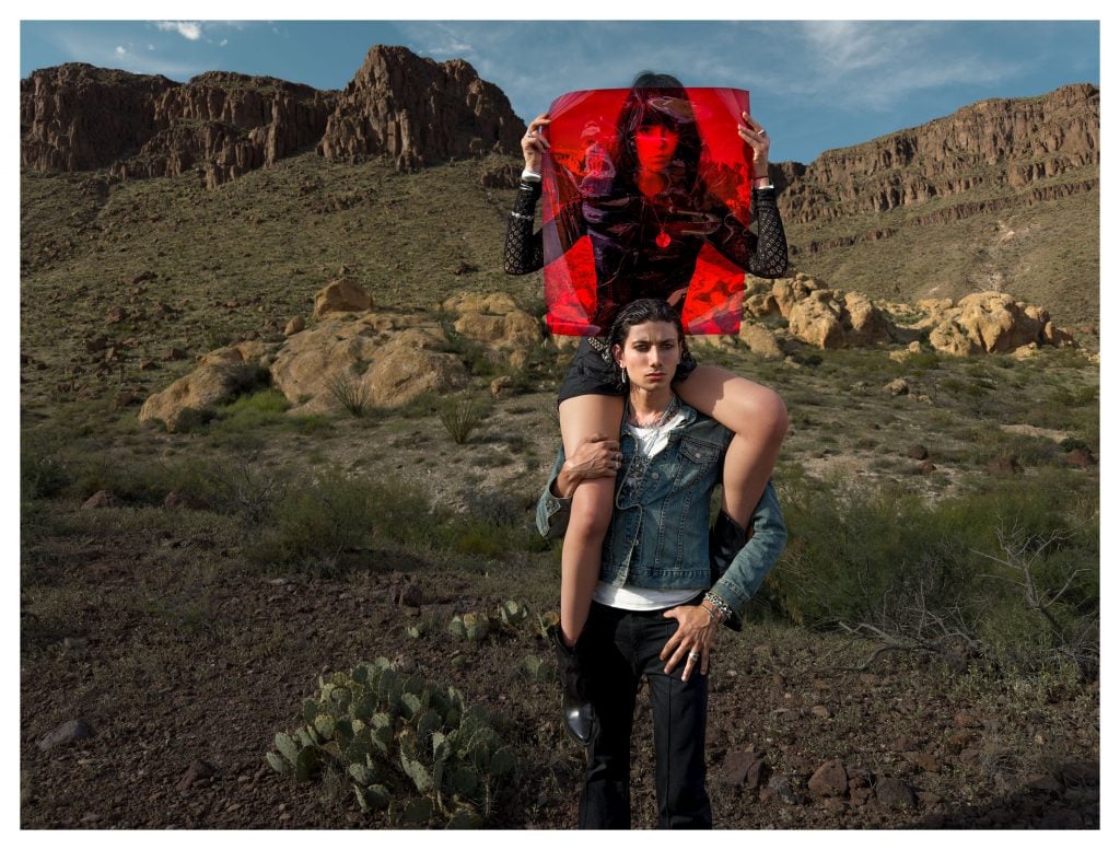 Portrait in desert of woman on man’s shoulders, holding red gel against mountainous backdrop.