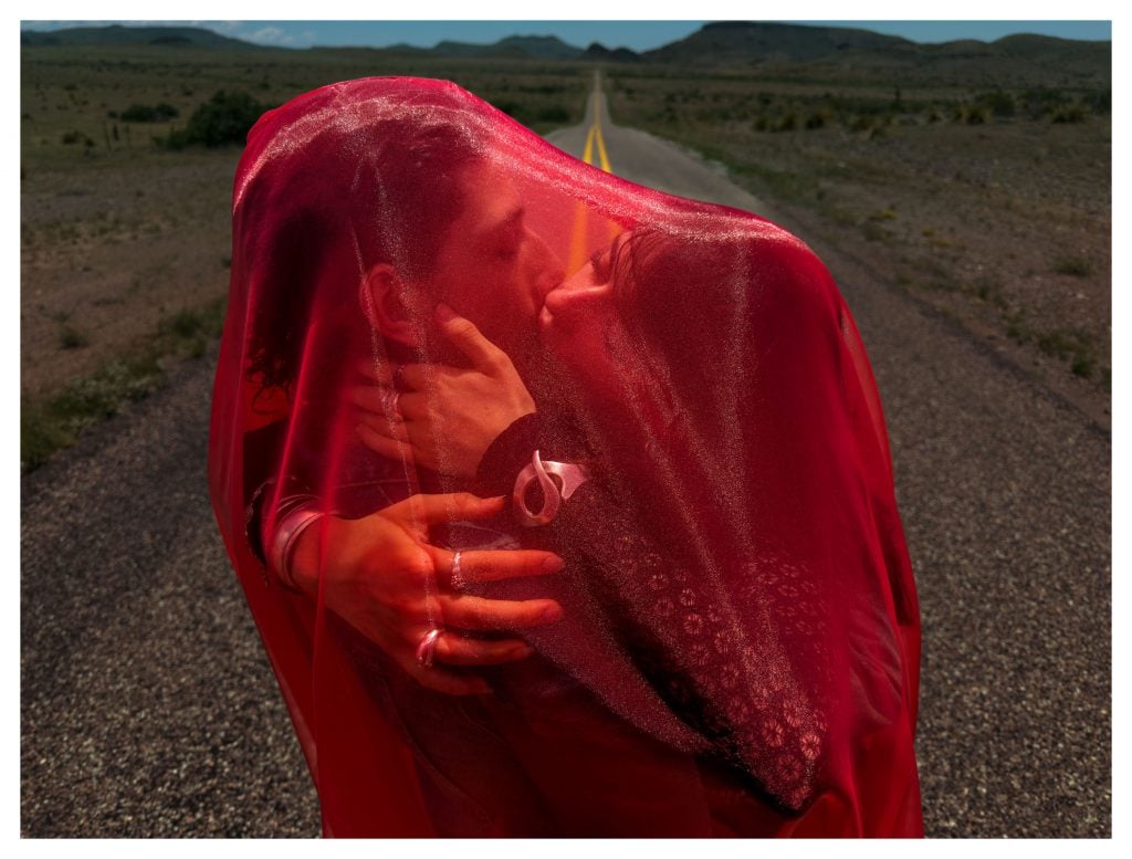 Couple kisses under translucent red fabric on open road, mountains faintly visible in background.
