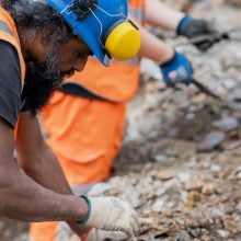 Archaeologist in blue hard hat and orange vest carefully excavating soil layers with precision tools.