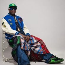 Virgil Abloh in green cap, sunglasses, blue varsity jacket, layered plaid skirts, and sneakers, seated casually.