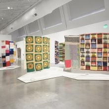 A photo of a bright gallery space displaying colorful quilts suspended from the ceiling, including designs with green and orange sunburst squares, patchwork ships, and geometric stripes.
