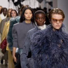 A line of models walking the runway in Bottega Veneta’s Spring/Summer 2026 finale. The leading model wears oversized sunglasses and a voluminous blue feathered coat, followed by others in sleek tailored looks and textured outerwear.