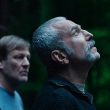 Two older men stand in a forest, one gazes upward intently, bathed in twilight.