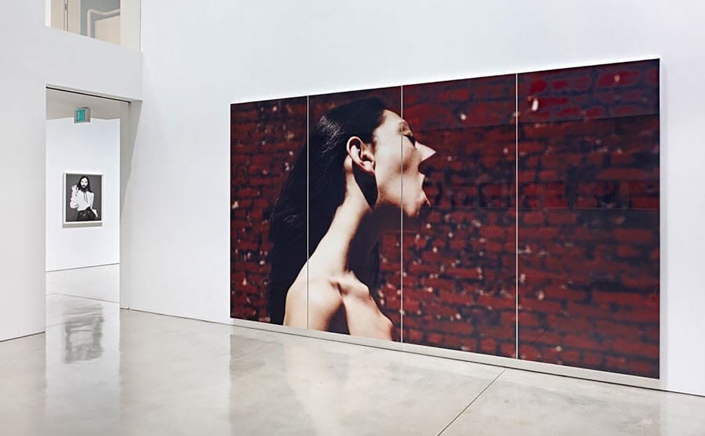 Large photograph of woman shouting against brick wall, displayed in white-walled gallery with polished floor.
