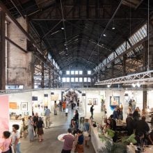 Wide view of the 2025 Atlanta Art Fair installation inside a large industrial hall with exposed beams and skylights, showing rows of gallery booths filled with artworks, colorful displays, and groups of visitors walking, conversing, and engaging with the art. A lively crowd gathers near the bar in the foreground, contributing to the energetic atmosphere.