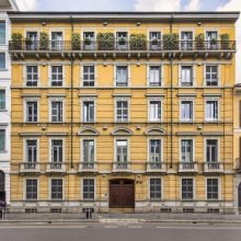 Exterior view of Mazzoleni Milan, a historic yellow building with white trim and tall windows, featuring a row of plants along the top floor balcony, situated between neighboring structures on a city street.
