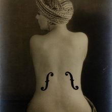 Black-and-white photograph of a woman with her back to the camera, wearing a turban, with violin-style f-holes painted onto her bare back.