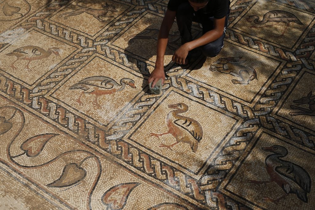 Person cleans ancient mosaic floor in Gaza, decorated with colorful birds, animals, and geometric patterns.