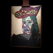 Painting by Pablo Picasso titled Buste de femme (Dora Maar) displayed on an easel, dramatically lit against a black background. The portrait features bold colors, geometric forms, and abstract floral motifs on the subject’s hat, characteristic of Picasso’s Cubist style.