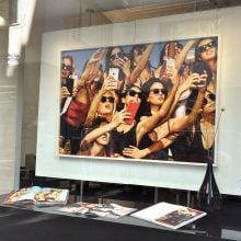 A gallery window display at Preiss Fine Arts in Beverly Hills featuring Tony Kelly’s 2018 artwork Model Nation, which depicts a group of women in sunglasses crowded together taking selfies with smartphones. Below the framed work, exhibition catalogs are open on a black surface, with a sleek black vase holding two yellow billy ball flowers completing the arrangement.